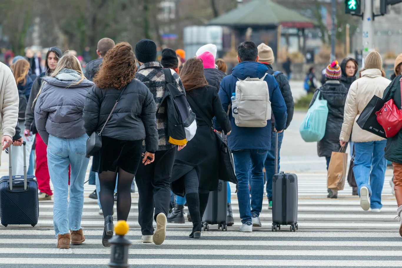 W końcu stycznia 2026 roku w urzędach pracy zarejestrowanych było 934,8 tys. bezrobotnych. To o 47 tys. więcej niż miesiąc wcześniej. Tłum osób w różnym wieku przechodzi przez przejście dla pieszych, wielu z nich ciągnie walizki. Część ludzi ubrana jest w ciepłe kurtki, w tle widoczne są drzewa oraz fragment budki lub kiosku.