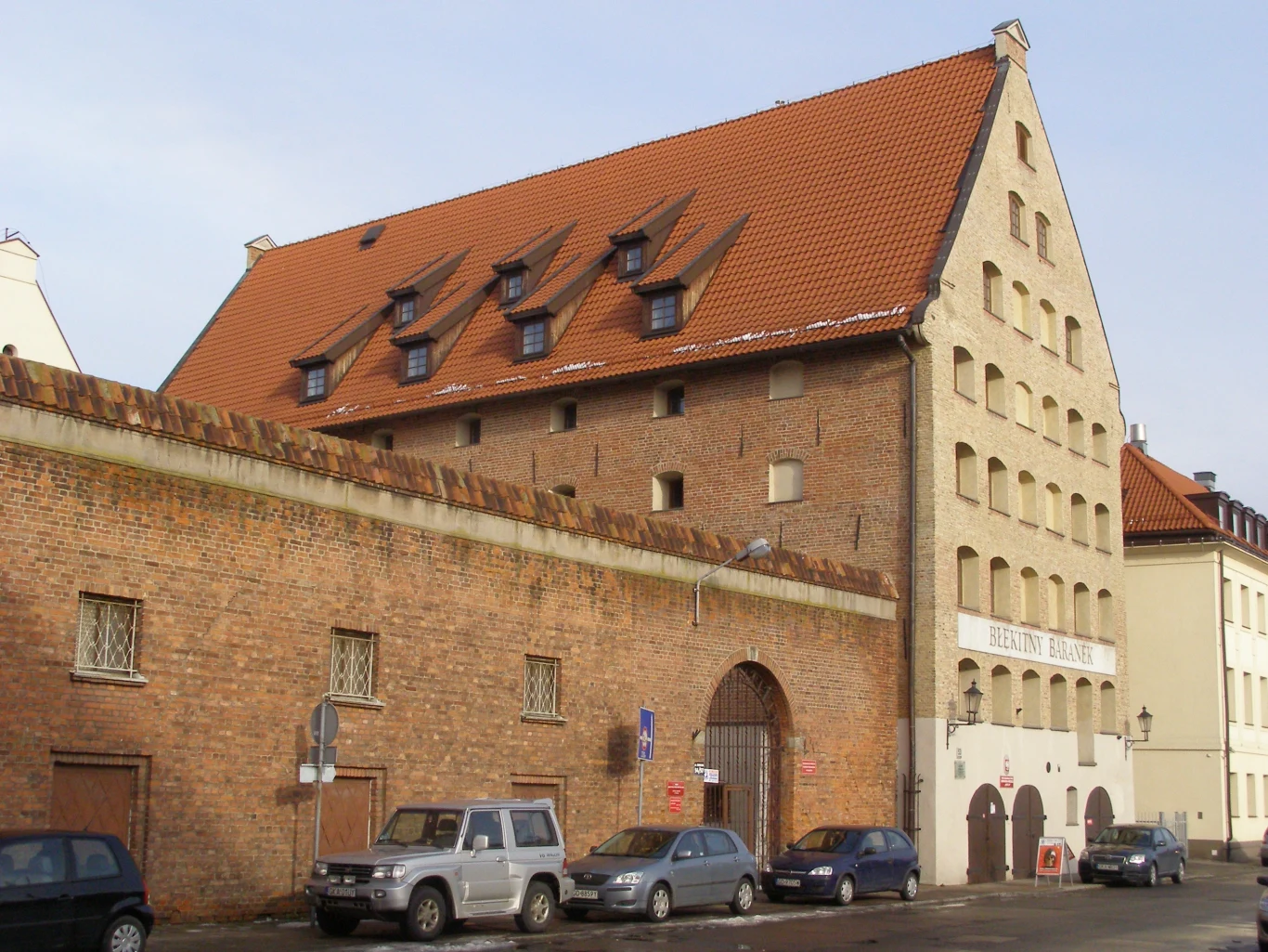 Budynek z czerwonym, spadzistym dachem i licznymi mansardowymi oknami, otoczony wysokim murem z cegły, za którym zaparkowane są samochody osobowe. Obiekt wygląda na zabytkowy i ma historyczną architekturę.
