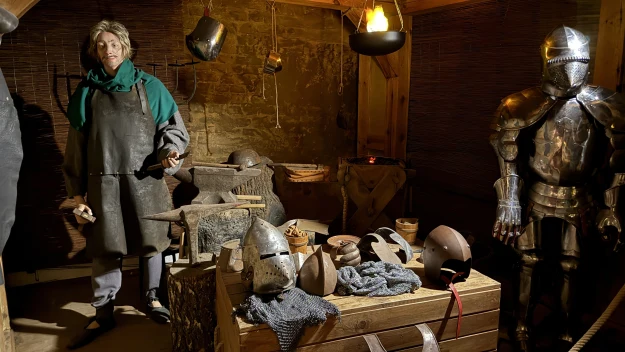Warsztat zbrojmistrza w średniowiecznym stylu, z manekinem ubraną w fartuch, paleniskiem, kowadłem i różnorodnymi elementami zbroi – hełmy oraz pełen pancerz rycerski, wszystko w drewnianym, klimatycznym wnętrzu o ciepłym oświetleniu.