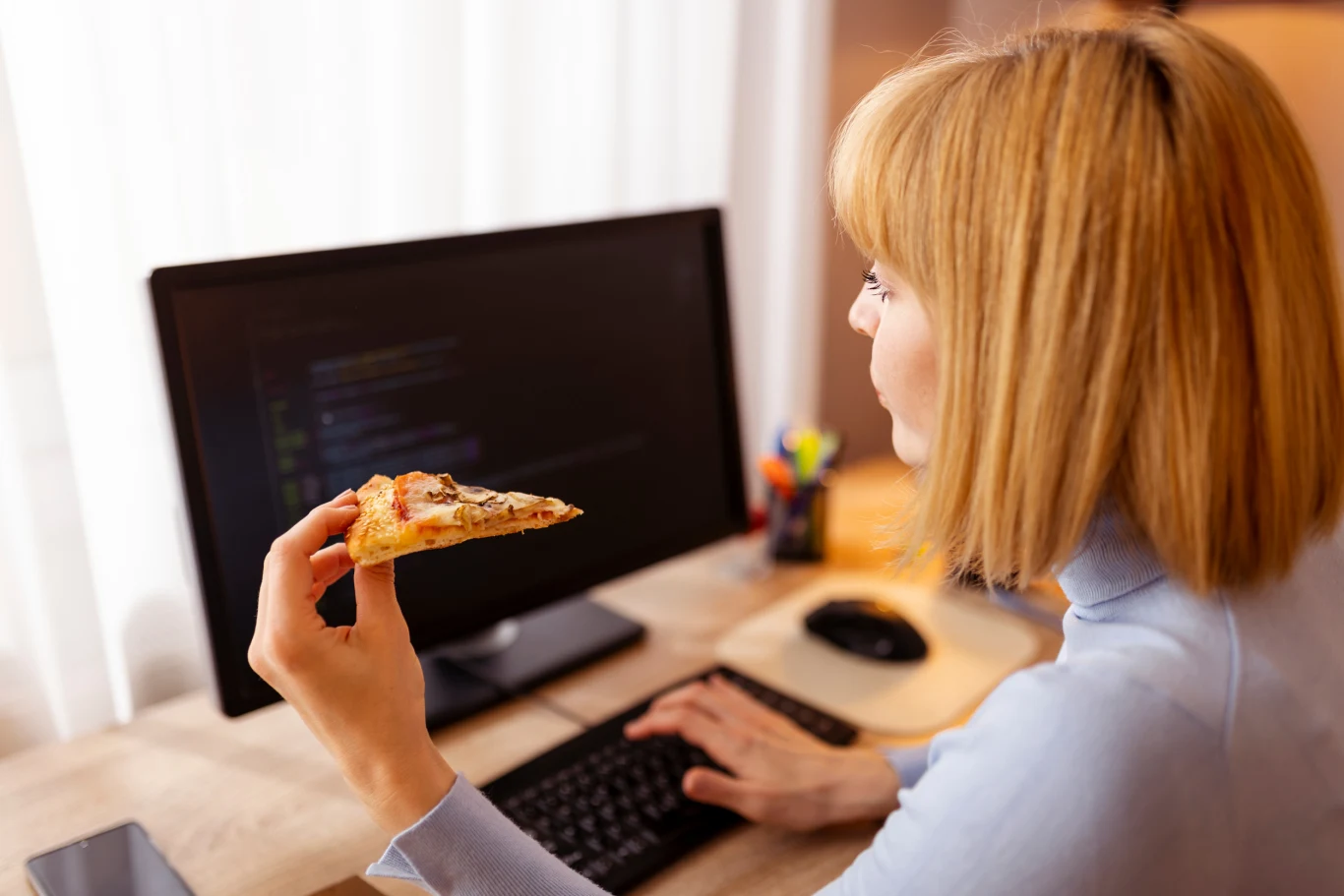 Kobieta pracuje przy komputerze, trzymając kawałek pizzy. Na monitorze widoczne są linie kodu, sugerujące programowanie lub pracę informatyczną. Na biurku znajduje się klawiatura, mysz i artykuły biurowe.