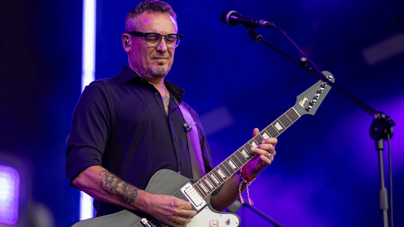 Hubert "Spięty" Dobaczewski Mężczyzna w okularach gra na gitarze elektrycznej, ubrany w ciemną koszulę, stoi na scenie oświetlonej fioletowym światłem.