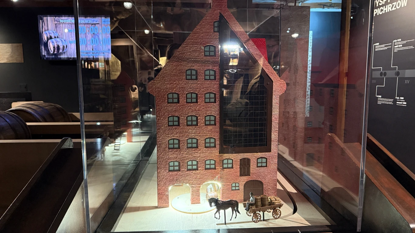 Model ceglanego budynku z czarnym dachem umieszczony za szkłem, przed nim stoi miniaturowy koń z wozem. W tle widoczne są elementy ekspozycji muzealnej, beczki oraz tablica informacyjna.