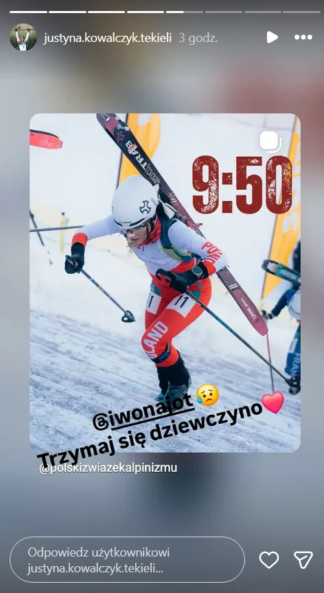 Zawodniczka w sportowym stroju narciarskim wspina się pod górę na śnieżnym stoku, trzymając narty na ramieniu. W tle widoczne są flagi organizatora oraz inni uczestnicy zawodów.