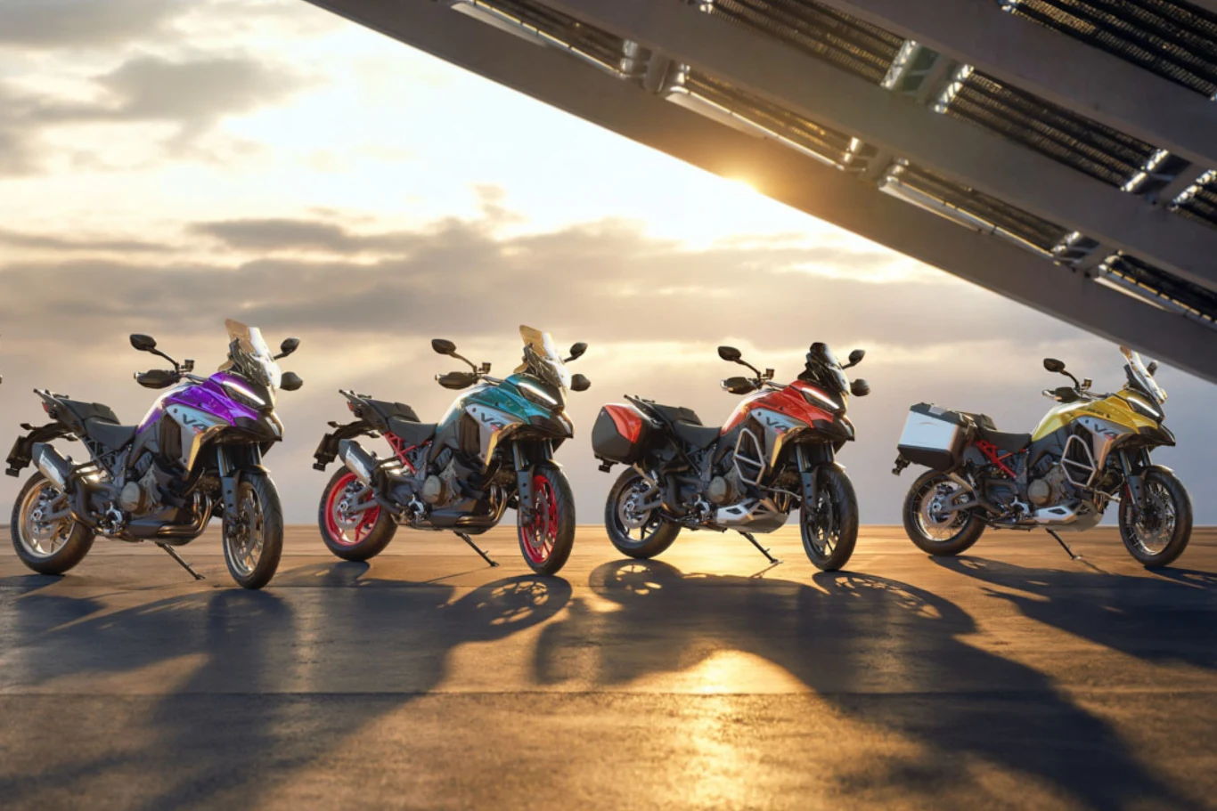 Cztery motocykle adventure ustawione w rzędzie na tle zachodzącego słońca, każdy w innym kolorze, pod nowoczesną konstrukcją architektoniczną.