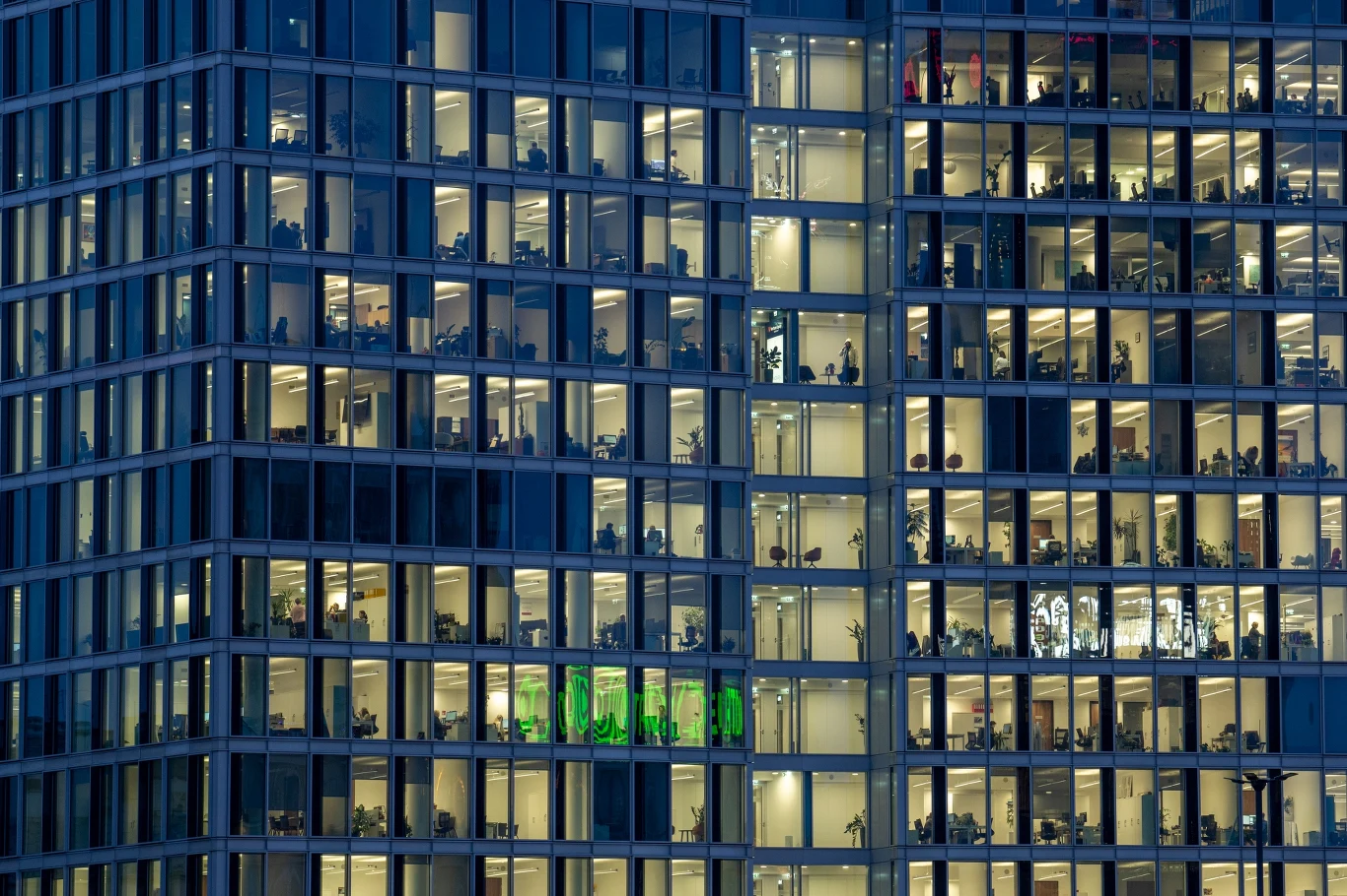Biurowiec; zdj. ilustracyjne Oświetlone biurowe piętra nowoczesnego wieżowca, widoczne przez wielkie okna, w których dostrzec można pracujących ludzi, biurka oraz elementy wyposażenia biurowego.