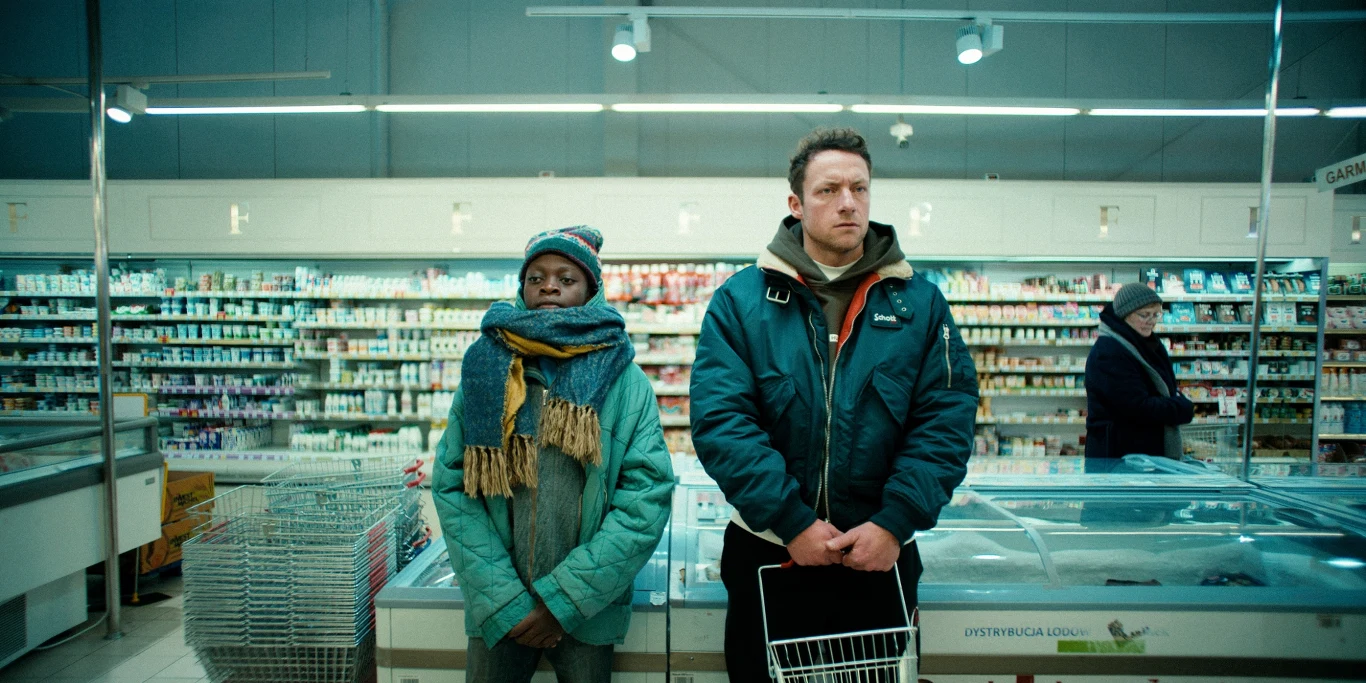 "Dobre chłopaki" Dwóch dorosłych stoi obok siebie przed zamrażarkami w supermarkecie, jedna osoba ubrana w kolorową zimową czapkę i szalik, druga w ciemnej kurtce, tło stanowią półki z nabiałem i innymi produktami spożywczymi oraz inna osoba robiąca zakupy.