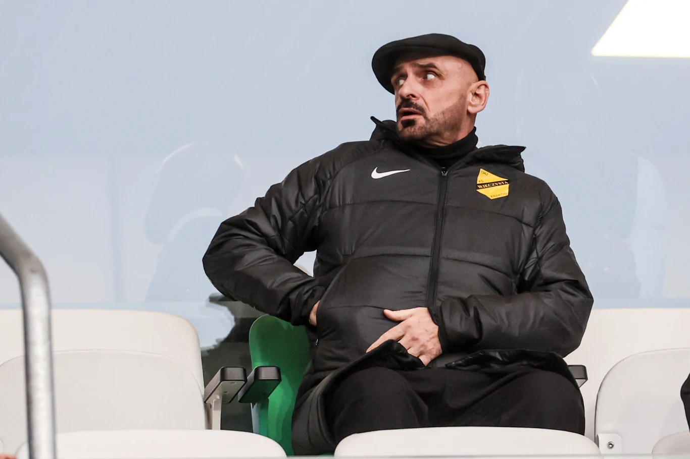 Mężczyzna w czarnej kurtce sportowej i czapce siedzi na trybunach, trzyma jedną rękę pod kurtką, patrząc w bok. Za nim rzędy pustych, białych foteli stadionowych.