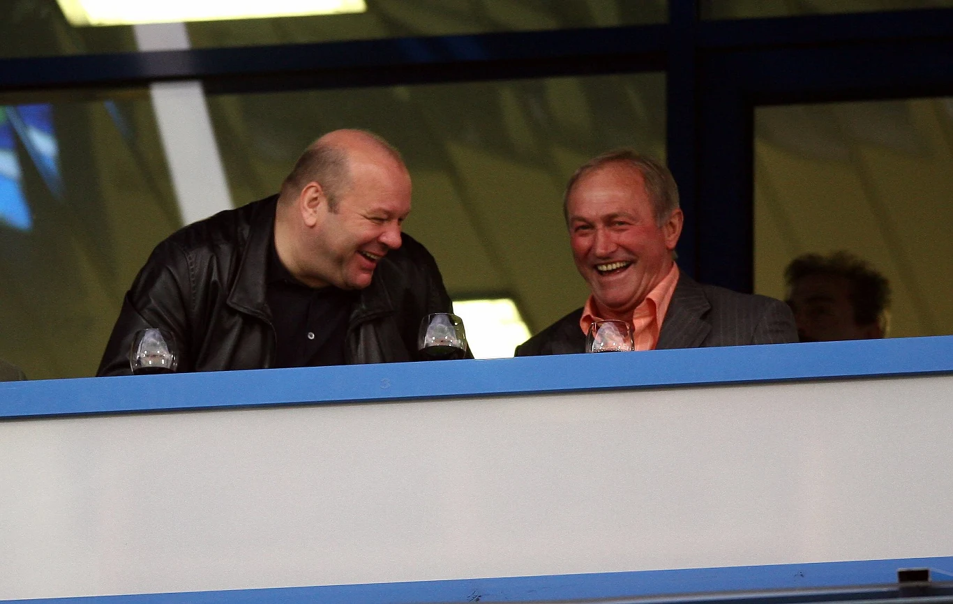 Dwóch starszych mężczyzn siedzi obok siebie na trybunie i śmieje się, obaj ubrani elegancko, atmosfera wydaje się radosna i swobodna.