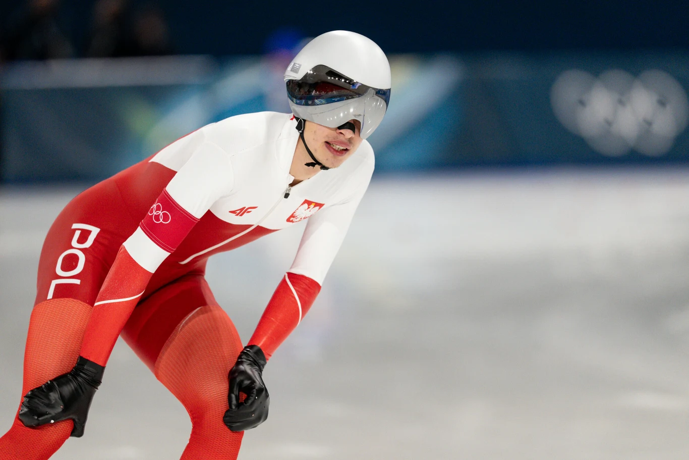 Władimir Semirunnij, 19.02.2026 r. Polski panczenista ubrany w biało-czerwony strój sportowy jedzie po lodowym torze podczas zawodów łyżwiarskich, w tle widoczne rozmyte logo olimpijskie.