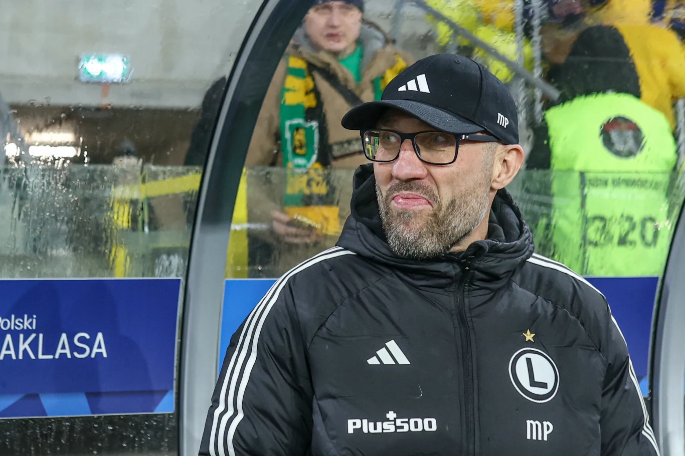 Trener Legii Warszawa ubrany w czarną kurtkę i czapkę z daszkiem stoi przy linii bocznej boiska, za nim widać kibiców w żółto-zielonych szalikach oraz stanowisko techniczne z tablicą z napisem PKO Bank Polski Ekstraklasa.