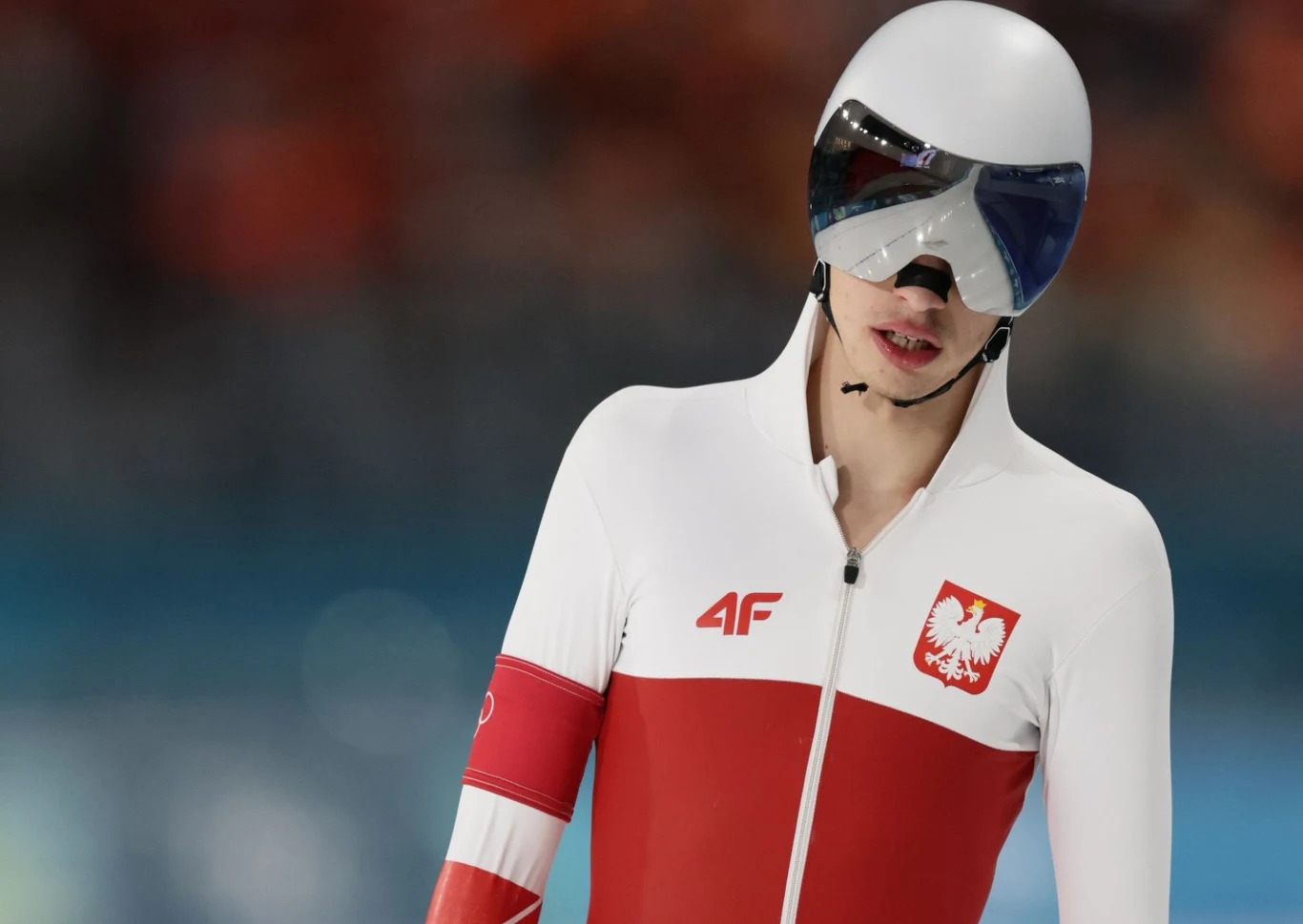 Łyżwiarz szybki w kombinezonie reprezentacji Polski, z charakterystycznym biało-czerwonym wzorem oraz godłem, w sportowym kasku i okularach na lodowisku.