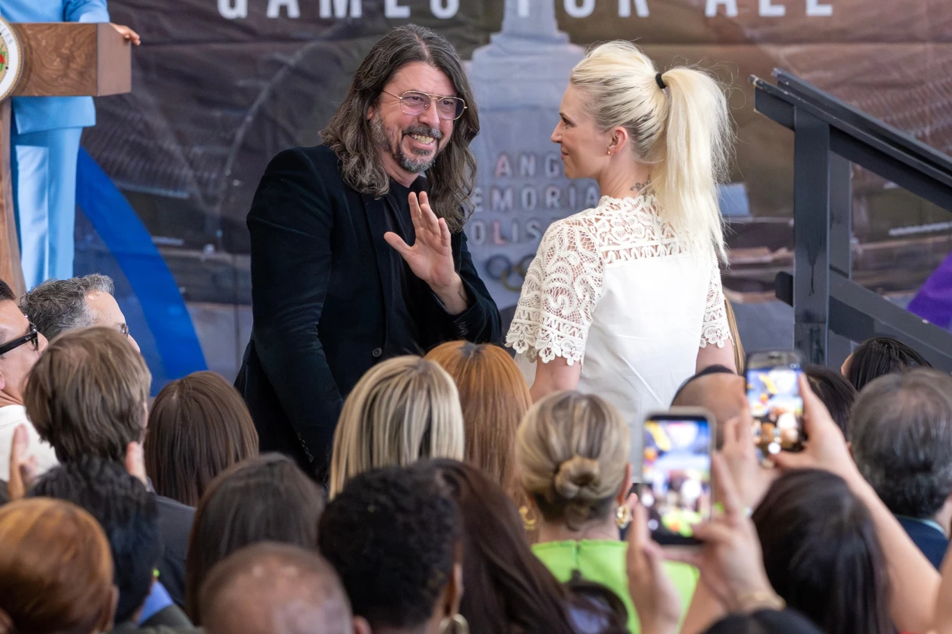 Dave Grohl jest liderem Foo Fighters, obok jego żona Jordyn Blum Mężczyzna z długimi włosami i brodą uśmiecha się i macha do tłumu podczas oficjalnego wydarzenia, obok niego stoi kobieta w białej bluzce, wokół zgromadziło się wiele osób, z których część robi zdjęcia telefonami.