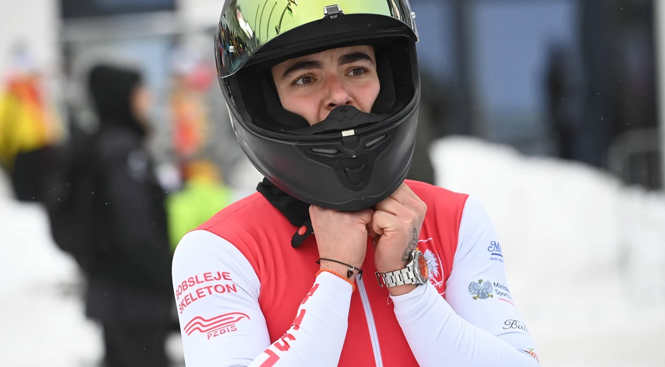 Linda Weiszewski Sportowiec w kasku z przyłbicą, ubrany w biało-czerwony strój z napisem bobsleje skeleton, przygotowuje się do startu na tle zimowego toru.