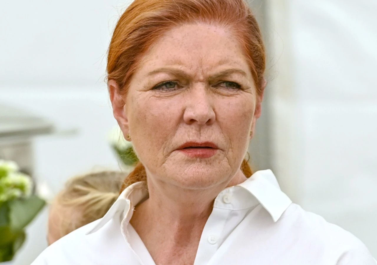 Katarzyna Dowbor Kobieta o rudych włosach spiętych do tyłu, ubrana w białą koszulę, z poważnym wyrazem twarzy na jasnym, neutralnym tle.