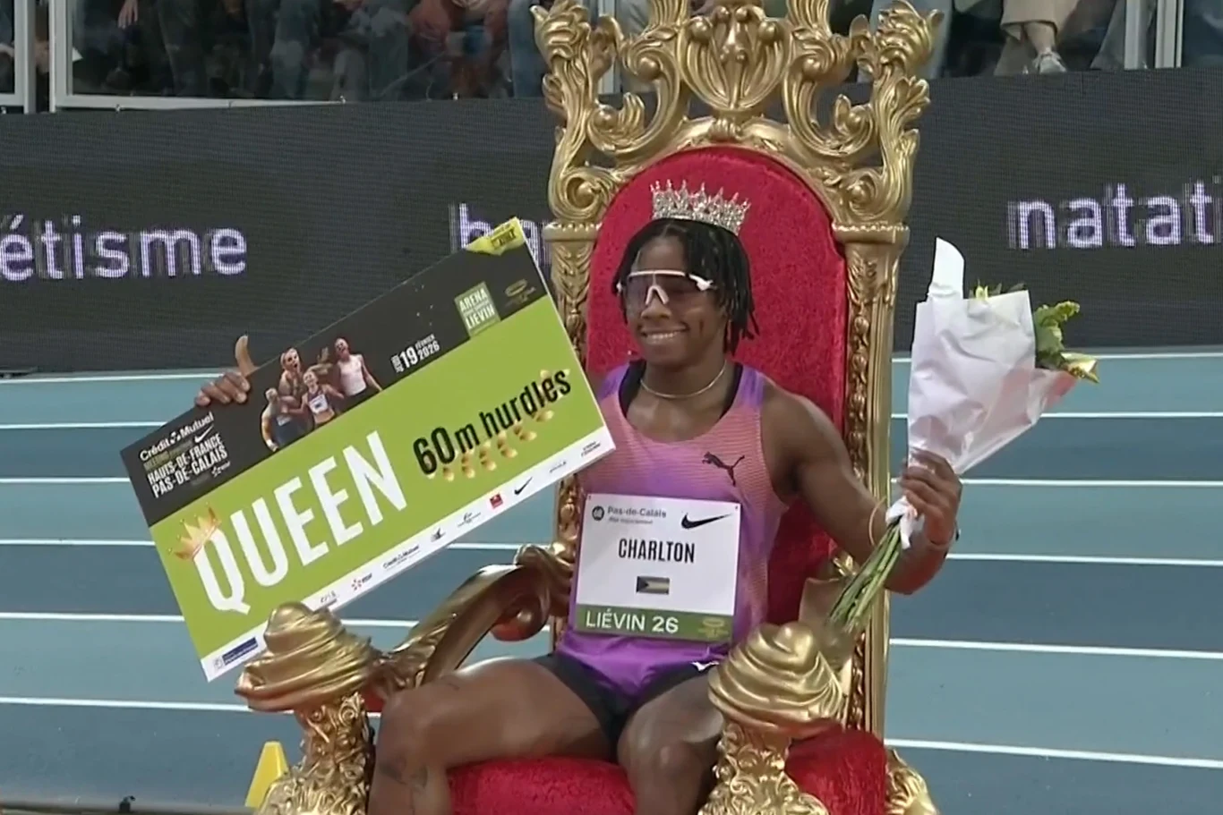 Zwyciężczyni biegu na 60 metrów przez płotki siedzi na złotym tronie, trzyma bukiet kwiatów oraz dużą tablicę z napisem Queen, na głowie ma koronę, a w tle widoczna jest hala sportowa.