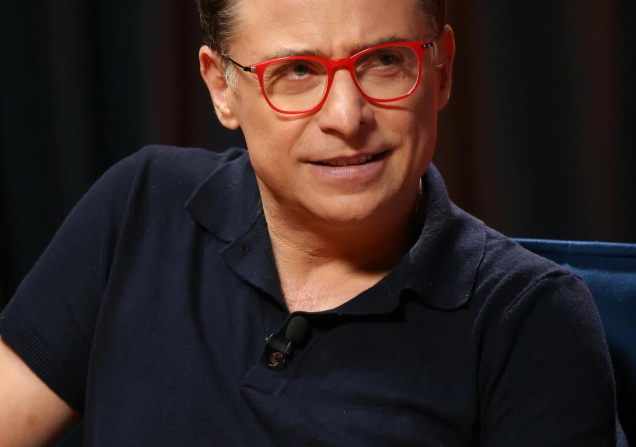 Kacper Kuszewski Mężczyzna w okularach w czerwonych oprawkach siedzi na tle ciemnych zasłon, ubrany w granatową koszulkę polo, z przypiętym mikrofonem do klapy.