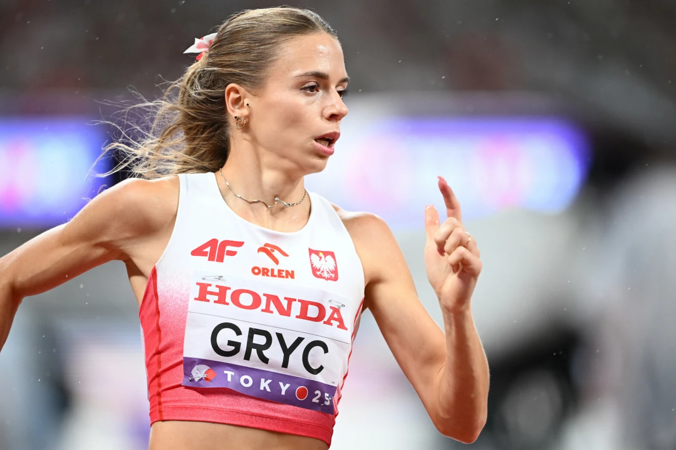 Lekkoatletka w biegu, ubrana w biało-czerwony strój z nazwiskiem na piersi oraz emblematem Polski, dynamicznie porusza się na stadionie.