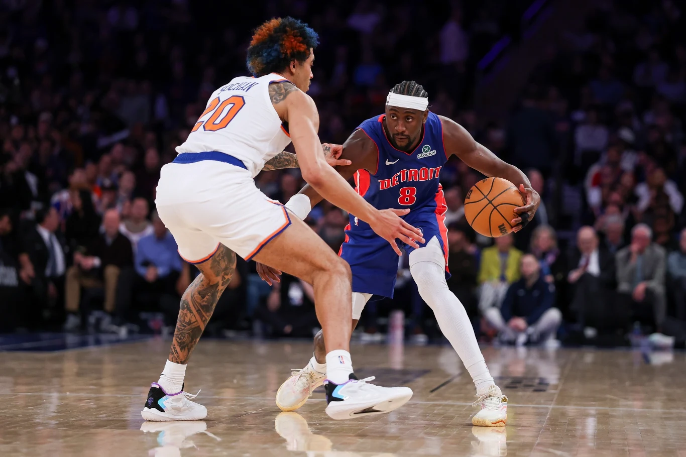 Dwóch koszykarzy walczy o piłkę podczas meczu, jeden z nich stara się minąć obrońcę na boisku pełnym kibiców. Intensywność rywalizacji podkreślają wyraziste pozy zawodników i skupienie na twarzach.