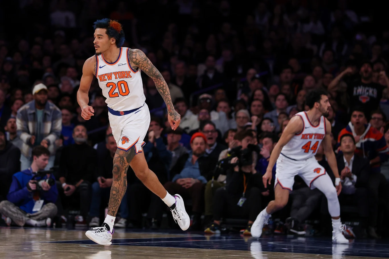 Dwóch koszykarzy drużyny New York Knicks w białych strojach biegnie po parkiecie podczas meczu. W tle liczna publiczność oraz osoby obsługujące wydarzenie.