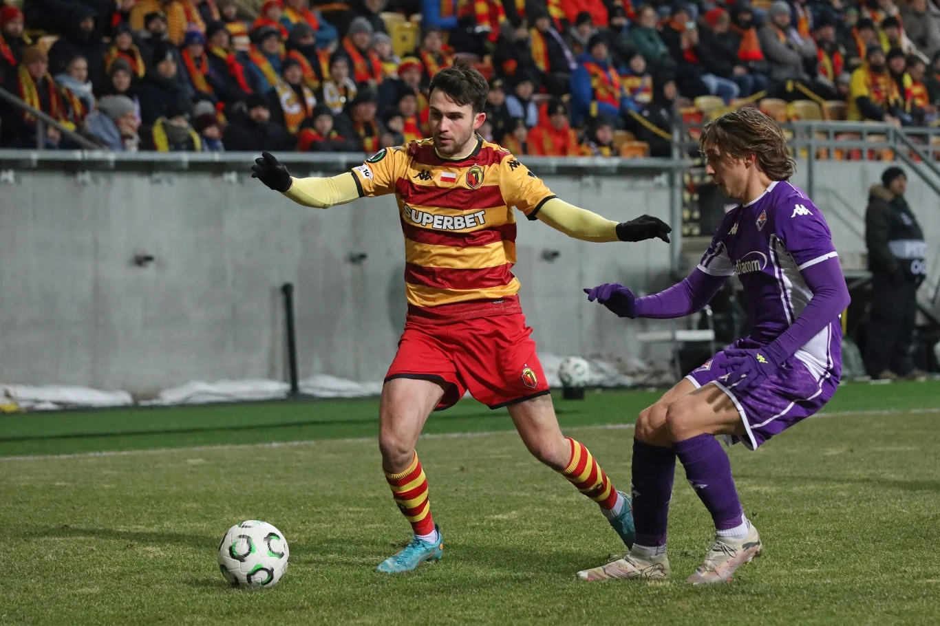 Dwóch piłkarzy rywalizuje o piłkę podczas meczu, jeden ubrany w żółto-czerwony strój, drugi w fioletowy. W tle kibice na trybunach.