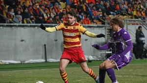 Dwóch piłkarzy rywalizuje o piłkę podczas meczu, jeden ubrany w żółto-czerwony strój, drugi w fioletowy. W tle kibice na trybunach.