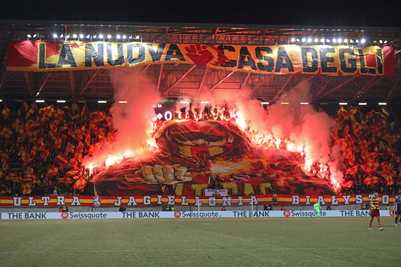 Kolorowa oprawa na stadionie piłkarskim z transparentem, dużą grafiką przedstawiającą postać oraz płonącymi racami, otoczona tłumem kibiców machających flagami w kolorach klubu.