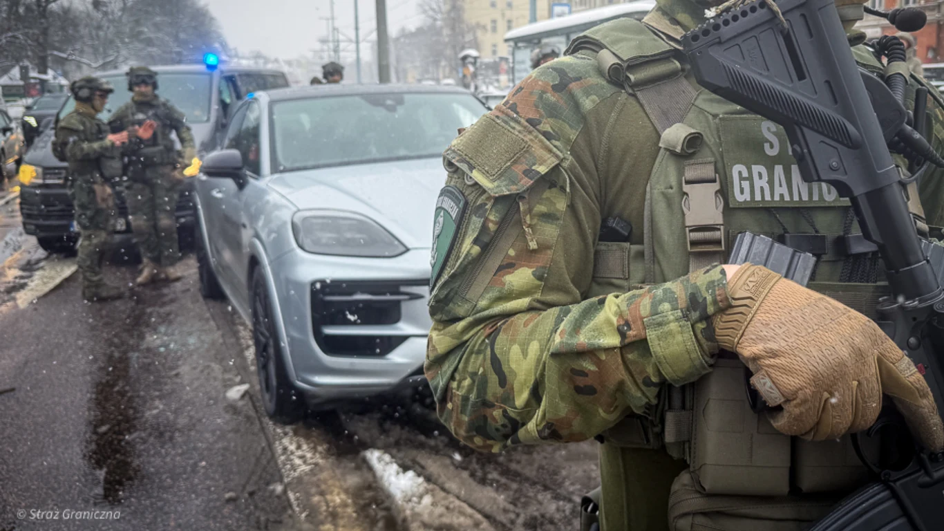 Straż Graniczna rozbiła grupę zajmującą się handlem ludźmi. Podczas pościgu funkcjonariusze użyli broni Umundurowani funkcjonariusze Straży Granicznej podejmują działania przy srebrnym samochodzie osobowym na ulicy w zimowych warunkach, z widoczną obecnością innych służb i mierzącymi bronią.
