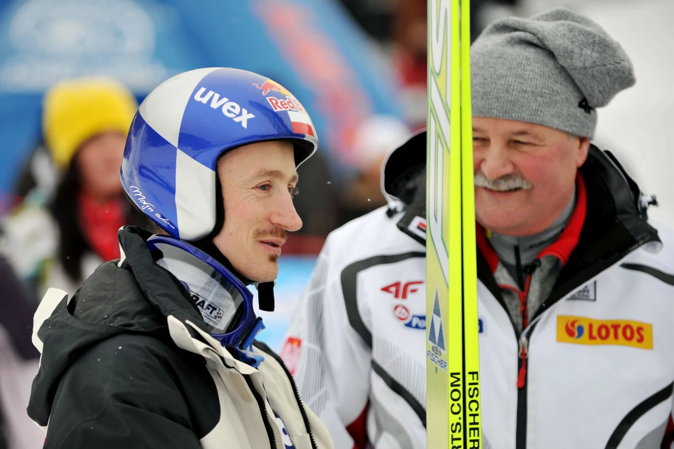 Dwóch mężczyzn ubranych w kombinezony sportowe podczas zawodów narciarskich, jeden z nich ma kask narciarski oraz trzyma narty z logo Fischer, w tle widoczne rozmyte sylwetki i elementy sponsoringu.