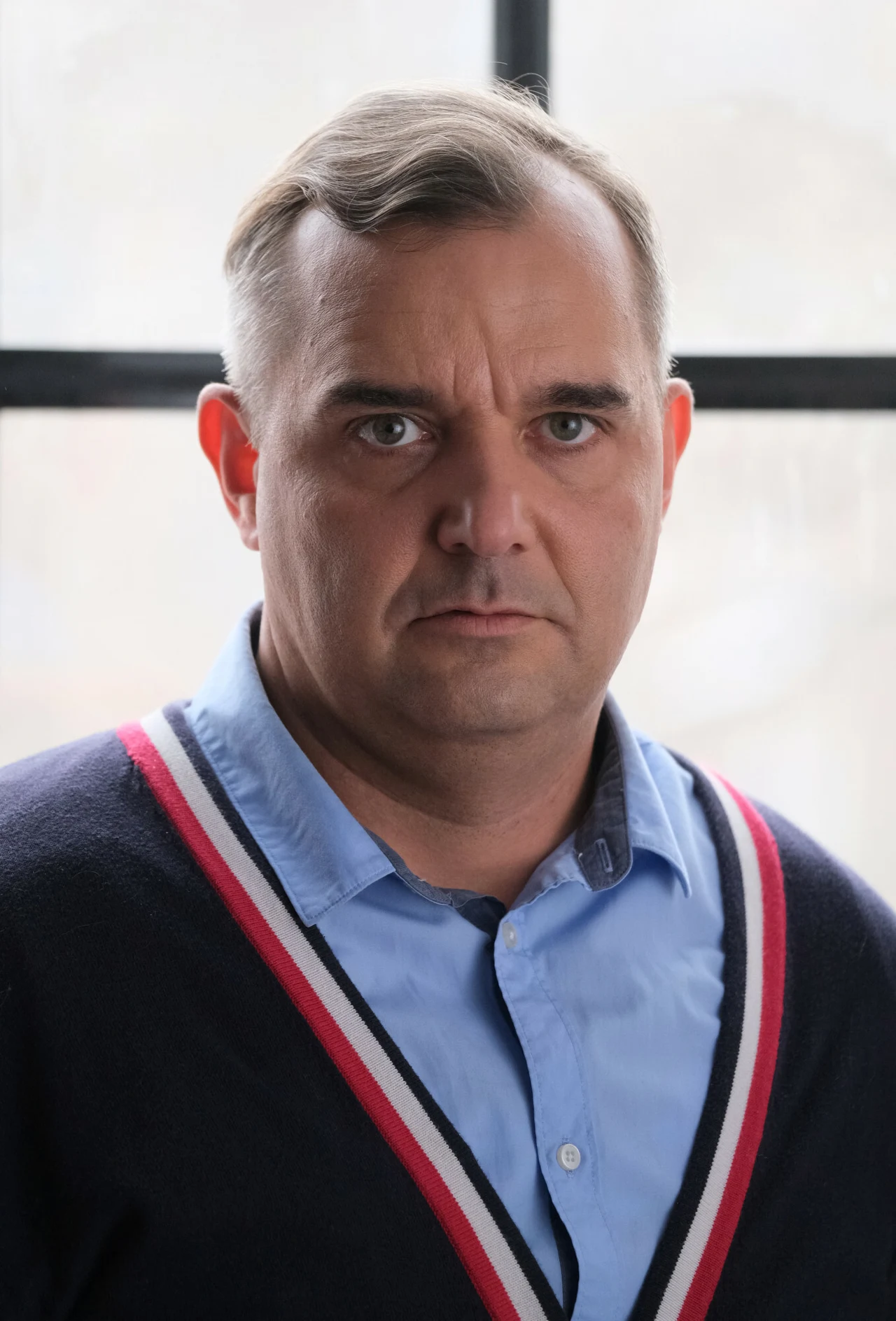 Mężczyzna o poważnym wyrazie twarzy stoi przed dużym oknem, ubrany w granatowy sweter z kolorowym paskiem i niebieską koszulę, patrzy prosto w obiektyw.