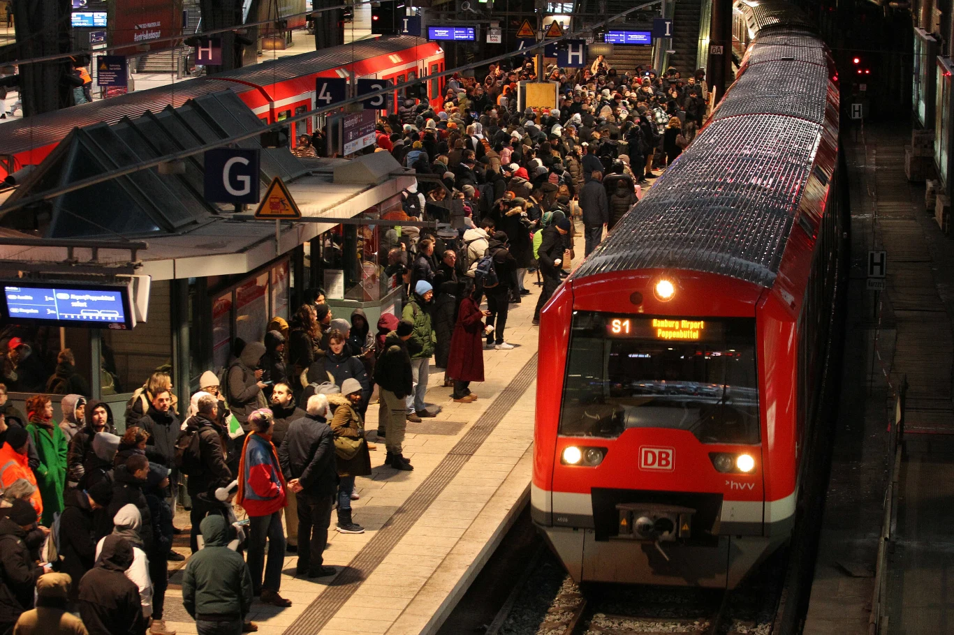Cyberatak na Deutsche Bahn (zdj. ilustracyjne) Zatłoczony peron kolejowy na dużej stacji, mnóstwo ludzi czeka na czerwony pociąg oznaczony logiem DB, wokół widoczne tablice informacyjne, oznaczenia peronów i jasne oświetlenie nocne.