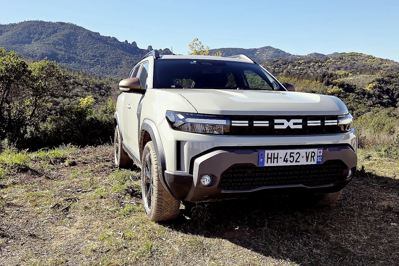 Dacia wprowadza do oferty nowy napęd Hyrid-150 4x4. Ma fabryczne LPG, automat i pali 7,3 l/100 km Samochód terenowy marki Dacia Duster o jasnym nadwoziu zaparkowany na otwartej przestrzeni z widokiem na górzysty krajobraz i roślinność śródziemnomorską w tle.