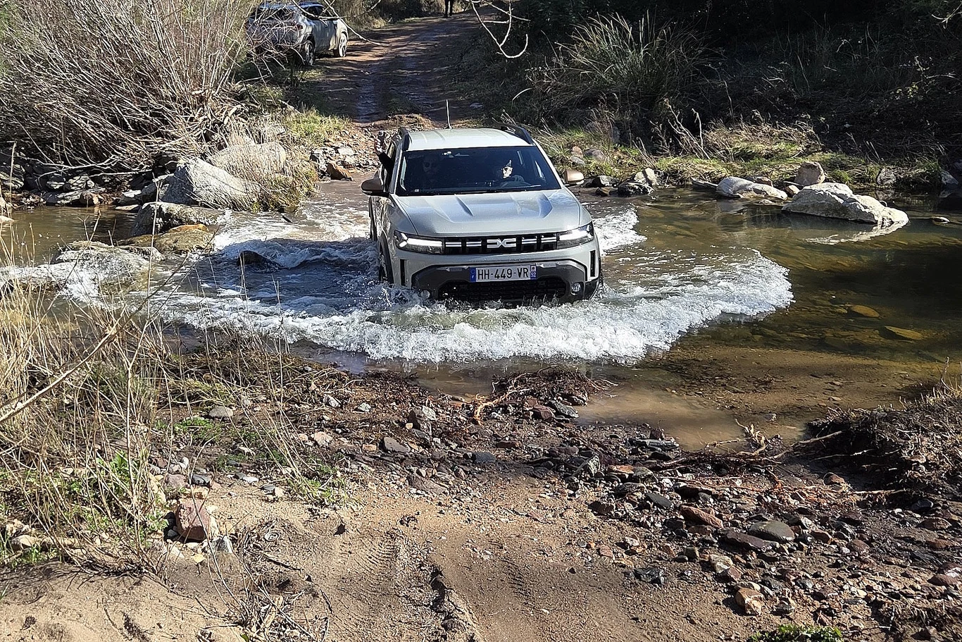 Dacia z napędem Hyrid-150 4x4 zaskakująco dobrze sprawuje się w terenie Samochód terenowy pokonuje płytką rzekę, wzburzając wodę, otoczony naturalną roślinnością typową dla terenu leśnego. W tle widoczny jest kolejny pojazd jadący po wyboistej drodze.
