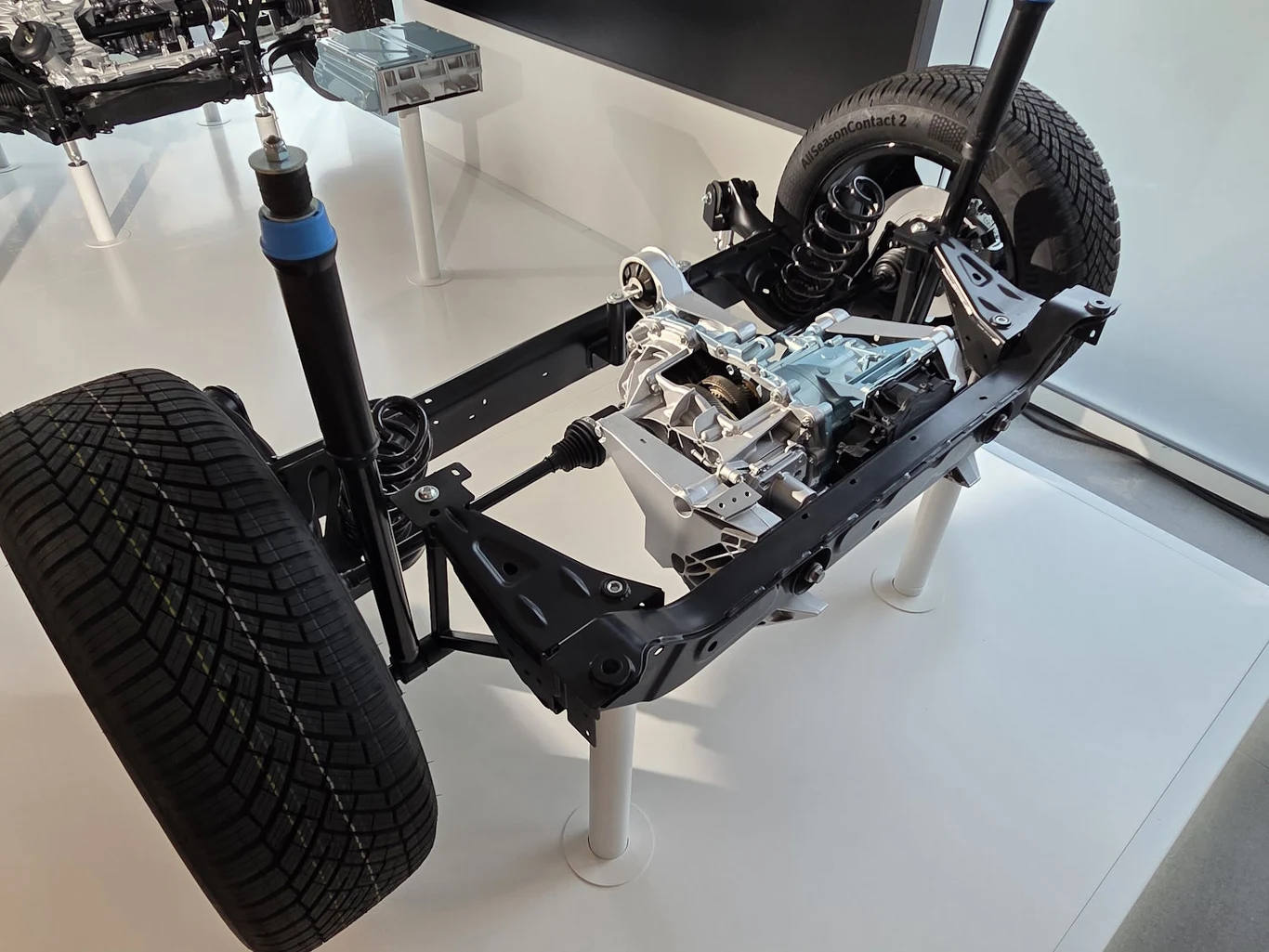 Zawieszenie samochodowe z widocznymi elementami układu jezdnego, na ekspozycji znajdują się koła, amortyzatory, sprężyny i ramy konstrukcyjne, całość ustawiona na białym podłożu laboratoryjnym.