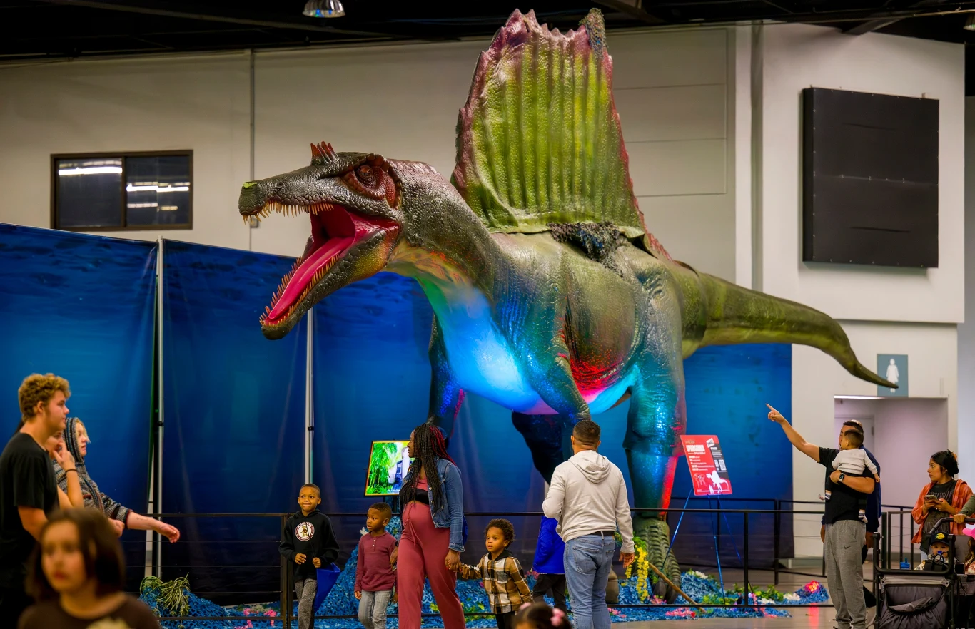 Duża makieta dinozaura typu Spinozaur z otwartą paszczą, przed którą stoją dzieci i dorośli zwiedzający wystawę. W tle niebieskie dekoracje imitujące podwodny świat. Ludzie podziwiają instalację, a niektóre osoby wykonują zdjęcia.