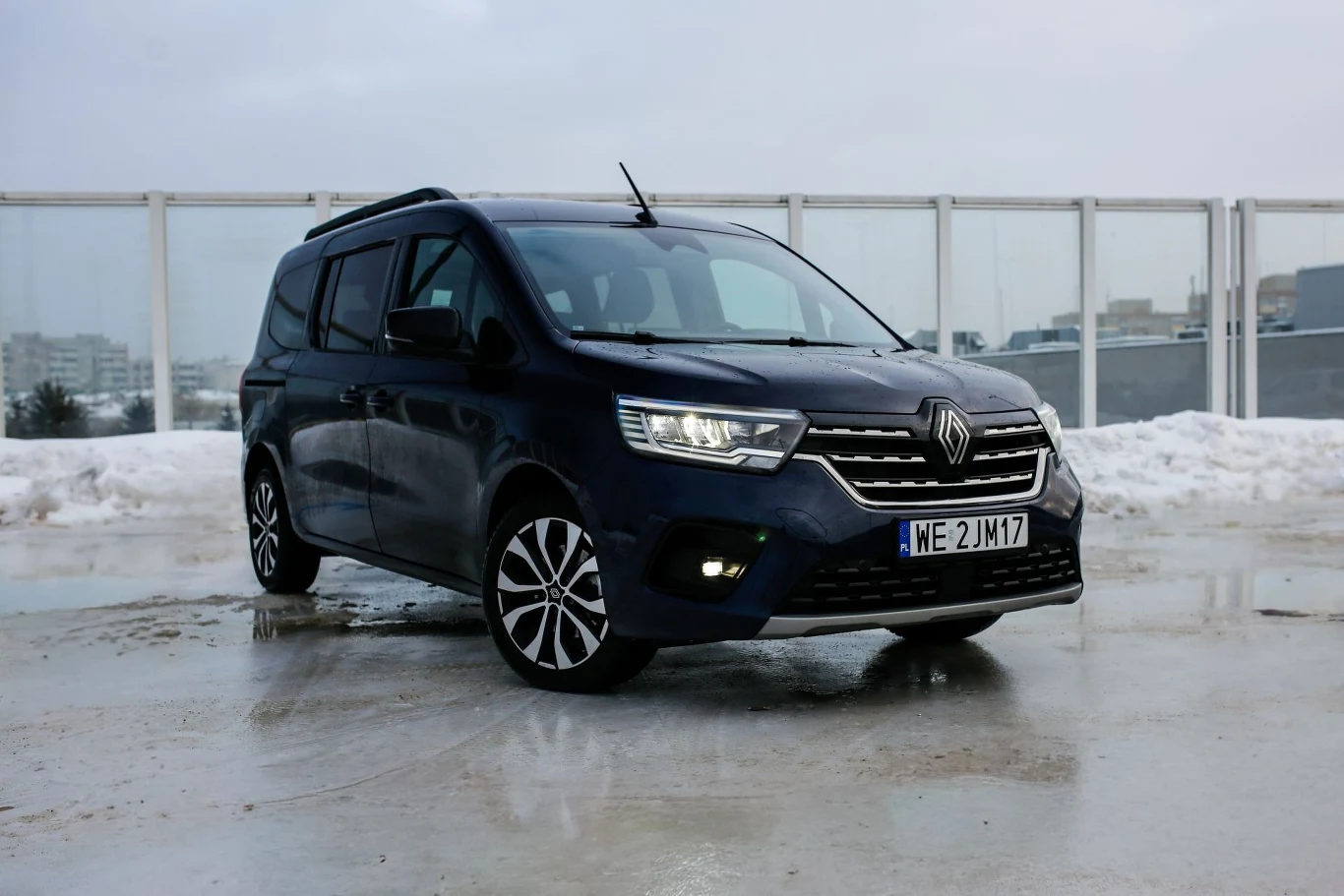 Renault Grand Kangoo to niemalże punkt odniesienia, jeśli chodzi o praktyczność w samochodach. Jedynym, ale bardzo znaczącym, minusem jest silnik. Czarny samochód Renault Kangoo zaparkowany na mokrej nawierzchni, w tle widoczna śnieżna sceneria i osiedle bloków mieszkalnych za przeszklonym ogrodzeniem.