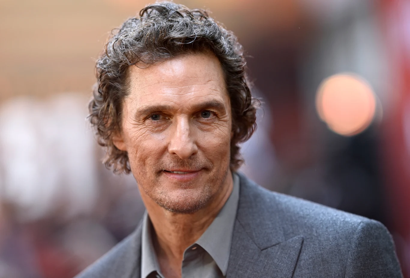 Matthew McConaughey w 2025 roku Mężczyzna w średnim wieku z lekko kręconymi, siwymi włosami i zarostem, ubrany w szary garnitur, patrzący bezpośrednio w obiektyw na rozmytym, jasnym tle.