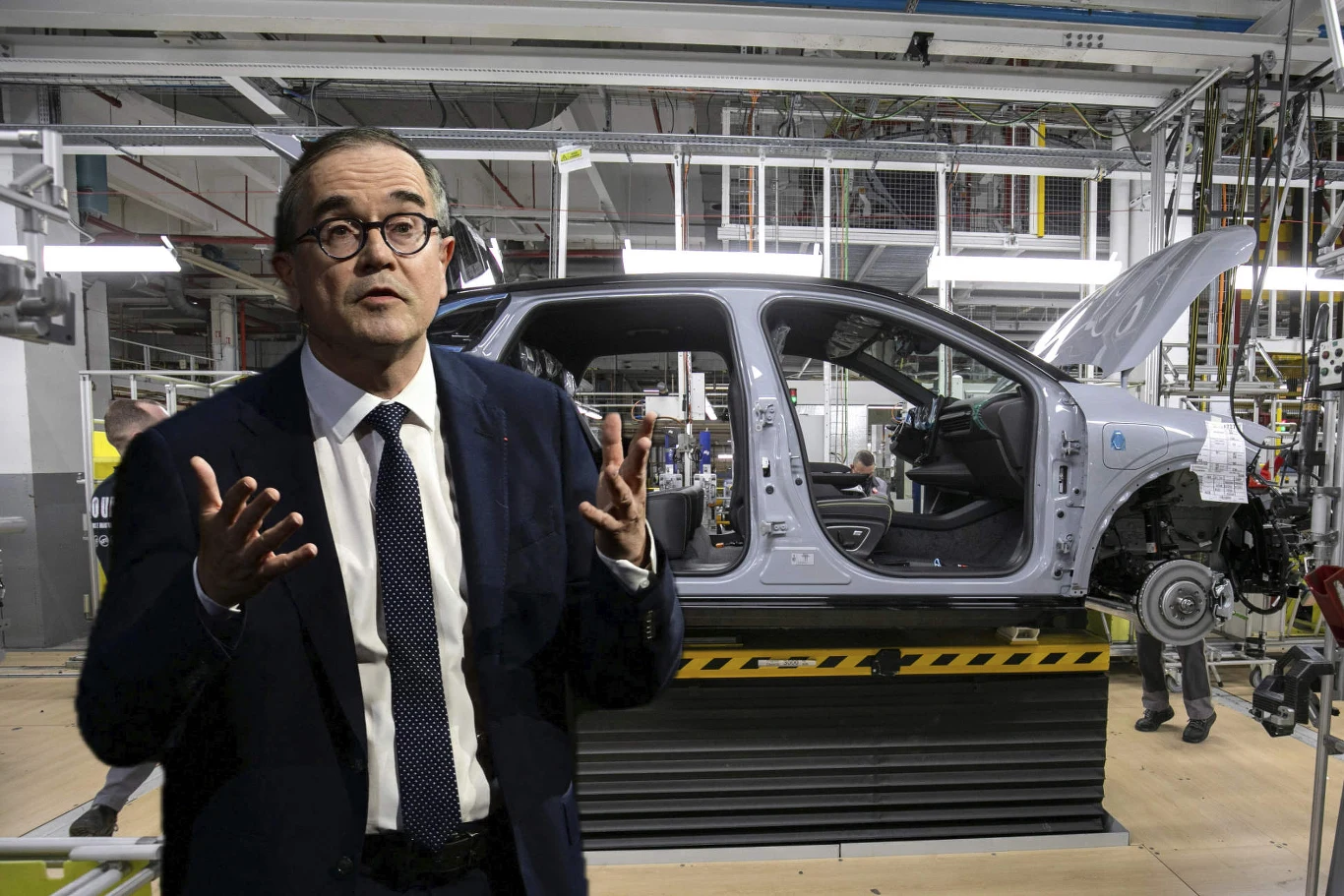 Mężczyzna w garniturze przemawia na tle linii produkcyjnej z montowanym nadwoziem samochodu w fabryce motoryzacyjnej.