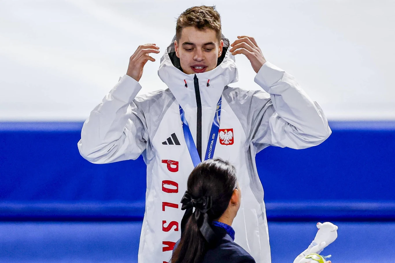 Sportowiec w białej kurtce z napisem 'Polska' oraz godłem, stojący na podium podczas ceremonii wręczenia medali, naprzeciwko osoby wręczającej nagrodę.