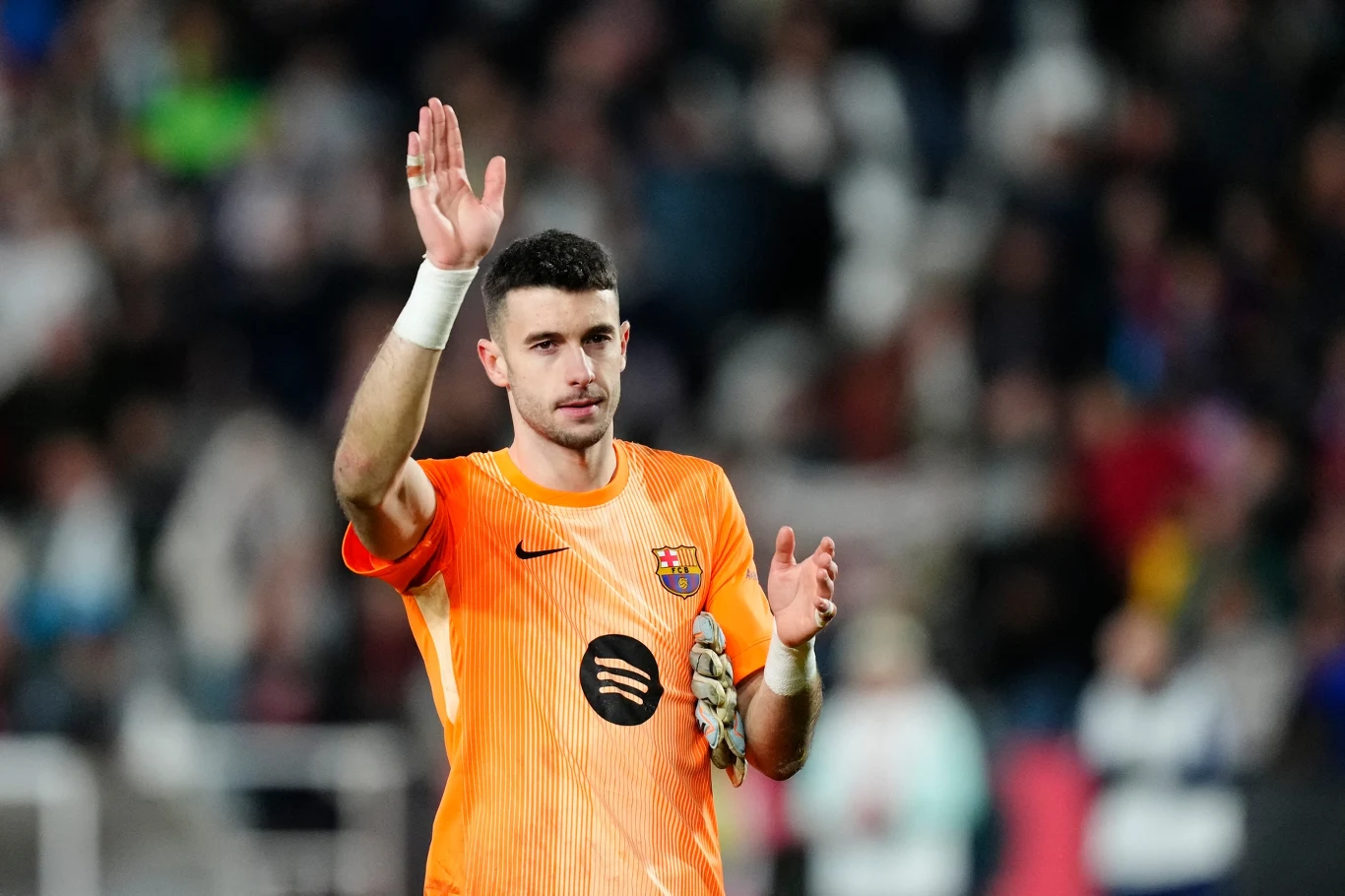 Młody mężczyzna w pomarańczowym stroju bramkarza FC Barcelony stoi na boisku i unosi rękę w geście pozdrowienia, na dłoni trzyma rękawice bramkarskie, w tle rozmyte sylwetki kibiców.