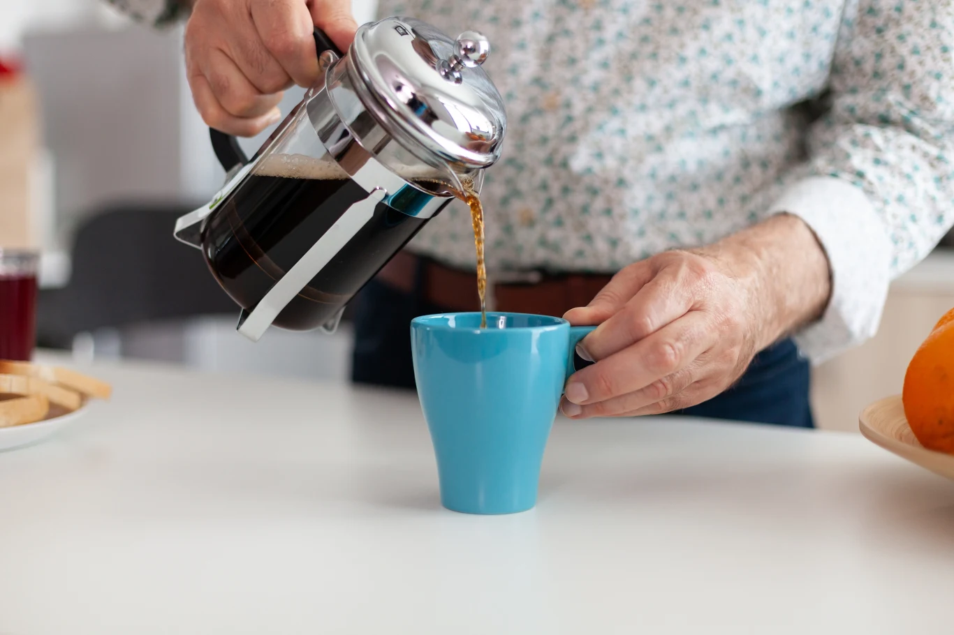 Osoba nalewa świeżo zaparzoną kawę z zaparzacza typu French press do niebieskiego kubka stojącego na białym blacie. W tle widoczne są fragmenty innych przedmiotów kuchennych, w tym miska z pomarańczami.