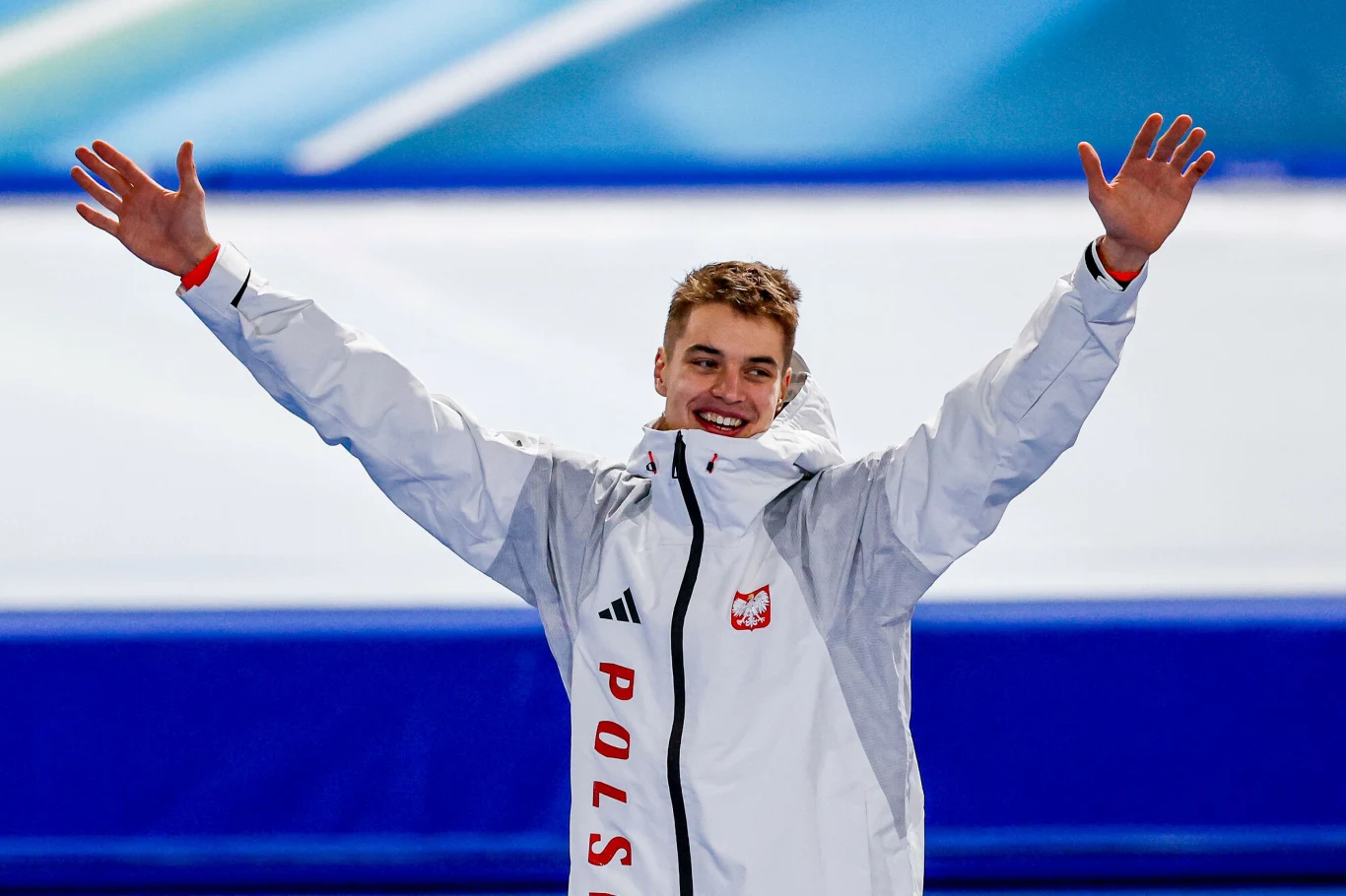 Władimir Semirunnij zdobył pierwszy dla Tomaszowa Mazowieckiego medal w łyżwarstwie szybkim Młody sportowiec w białej kurtce z napisem Polska i godłem narodowym stoi z szeroko uniesionymi rękami, uśmiechnięty, na tle niebieskiej areny sportowej.