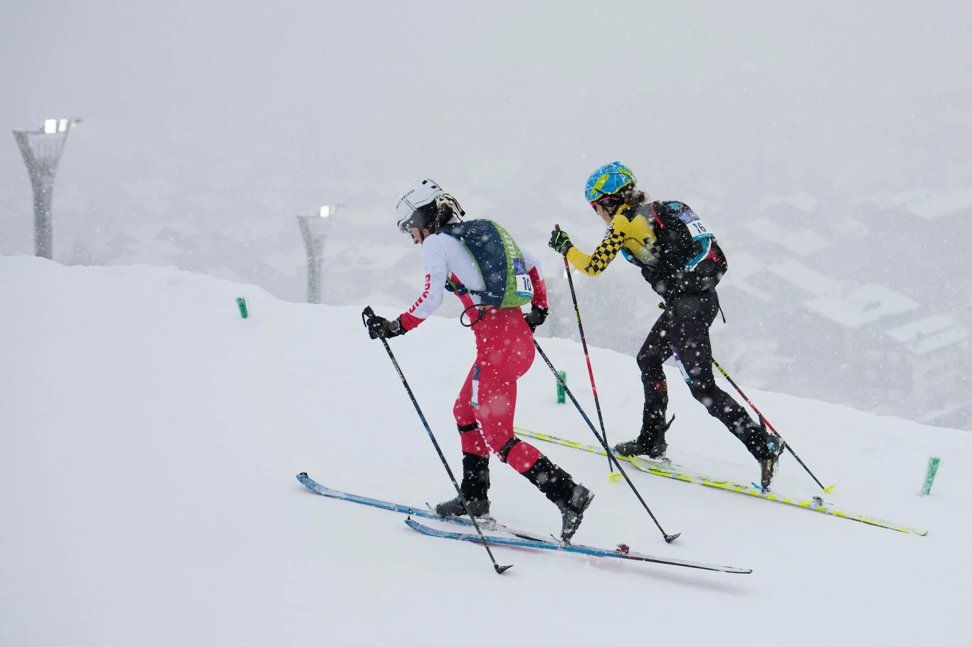 Dwóch narciarzy biegowych idzie w górę zaśnieżonego stoku podczas intensywnych opadów śniegu, ubrani w sportowe kombinezony i kaski z numerami startowymi, wokół panują zimowe warunki.