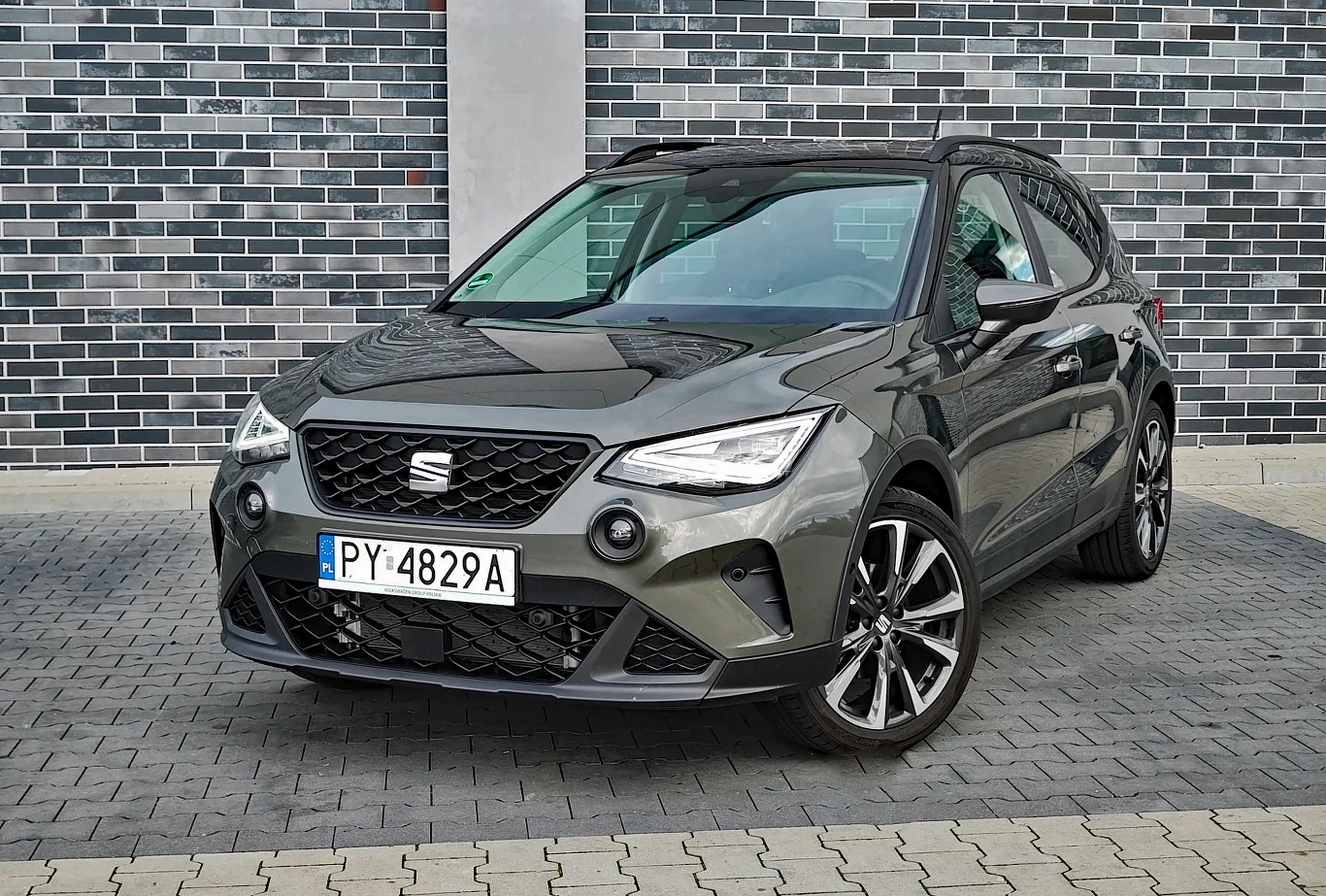 Seat Arona ma solidny staż rynkowy, ale stylistycznie wygląda świeżo Szary samochód osobowy marki Seat Arona stoi na brukowanej nawierzchni na tle ściany z szarych cegieł, widoczny z przodu i z boku.