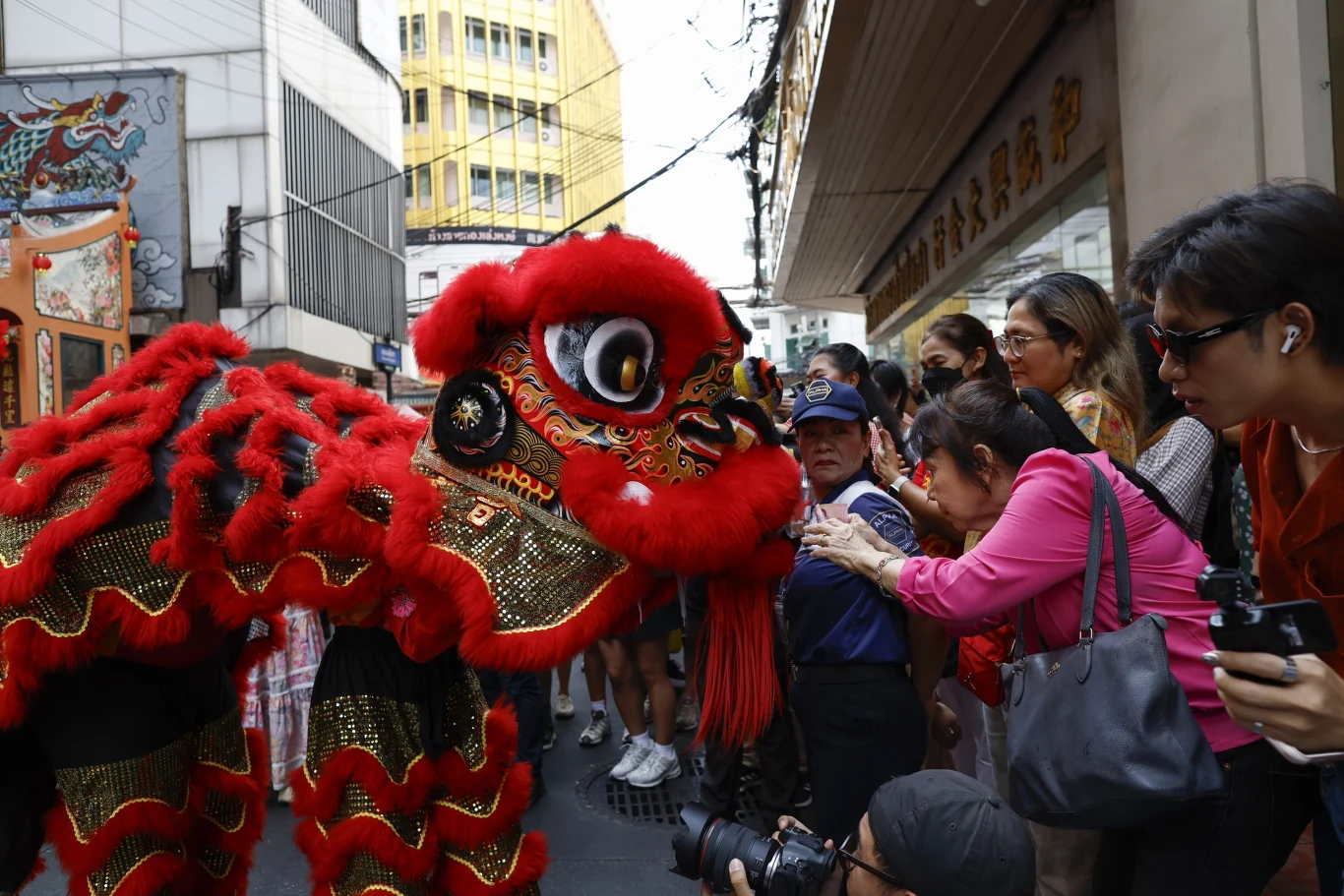 Bangkok. Uczestnicy parady z okazji księżycowego nowego roku Czerwono-czarny kostium lwa podczas tradycyjnego chińskiego tańca na ulicy, wokół zebrała się grupa ludzi fotografujących i nagrywających występ. W tle widoczne miejskie budynki i sklep.