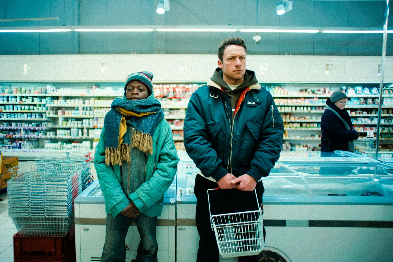 Samson Owino Ochieng i Konrad Eleryk na planie "Dobrych chłopaków" Dwóch mężczyzn stoi obok siebie przed lodówką w supermarkecie, jeden ubrany w ciepłą kurtkę i czapkę, trzyma pusty koszyk na zakupy, drugi ubrany w turkusową kurtkę i szalik, również w czapce zimowej. W tle regały chłodnicze z nabiałem oraz stojąca z b...