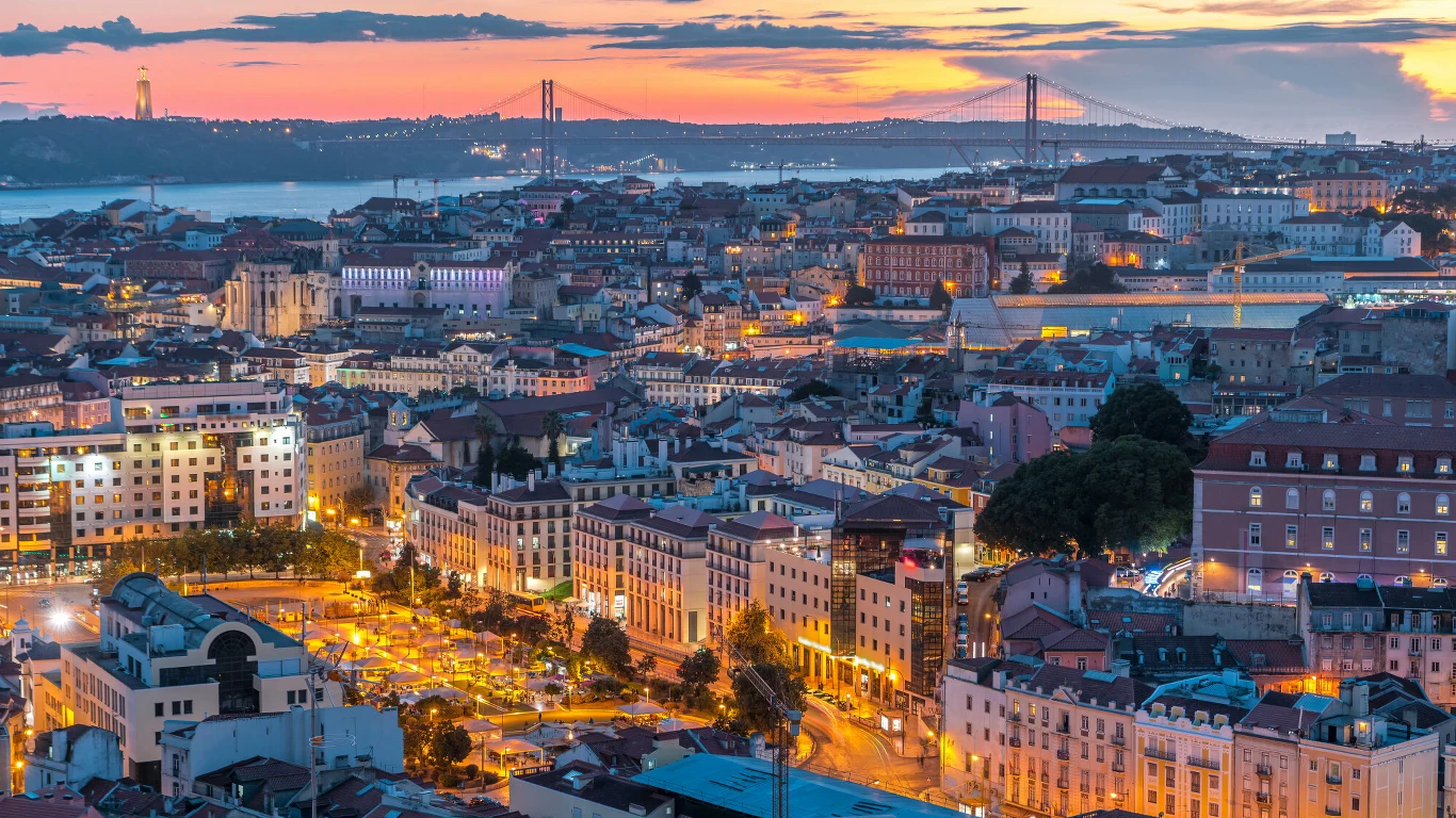 Lizbona jesienią zachwyca jeszcze bardziej Panorama miasta z licznymi kolorowymi budynkami, mostem rozciągającym się nad rzeką na drugim planie oraz zachodem słońca rozświetlającym niebo. Ulice i place są oświetlone światłami miasta, tworząc przytulną atmosferę.
