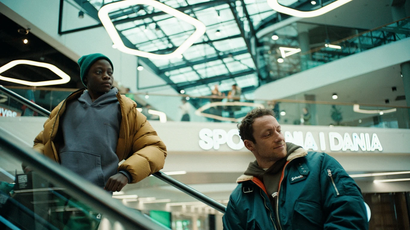 Samson Owino Ochieng to odkrycie "Dobrych chłopaków" Dwóch mężczyzn w zimowych kurtkach jedzie ruchomymi schodami w nowoczesnym centrum handlowym pod przeszklonym dachem, w tle widoczne neonowe napisy i geometryczne lampy.