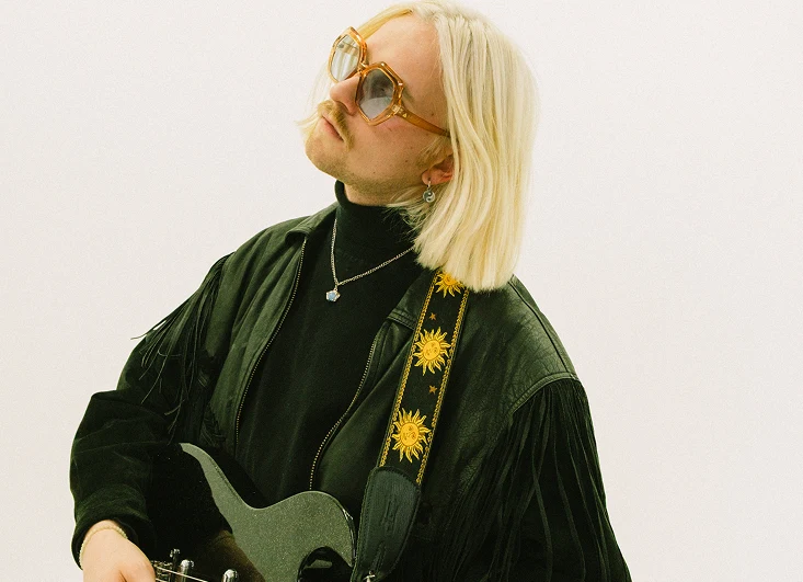 Dolphin Love wystąpi na NEXT FEST 2026 Mężczyzna z długimi blond włosami i w dużych okularach przeciwsłonecznych, ubrany w czarną skórzaną kurtkę z frędzlami i czarny golf, grający na elektrycznej gitarze, pasek z motywem słońca.