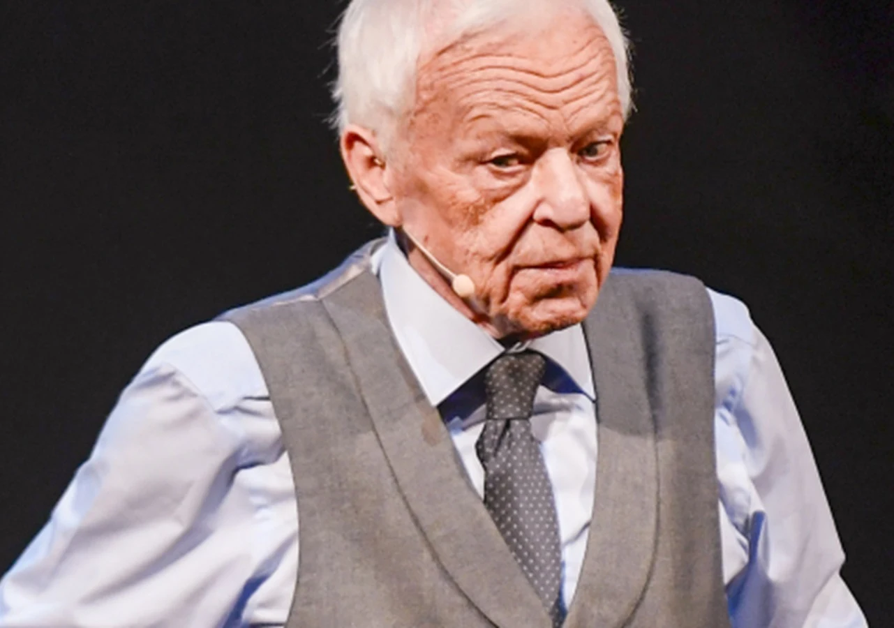 Marian Opania Starszy mężczyzna w eleganckim stroju, mikrofon na uchu, skupiony wyraz twarzy, ciemne tło podkreślające sylwetkę.