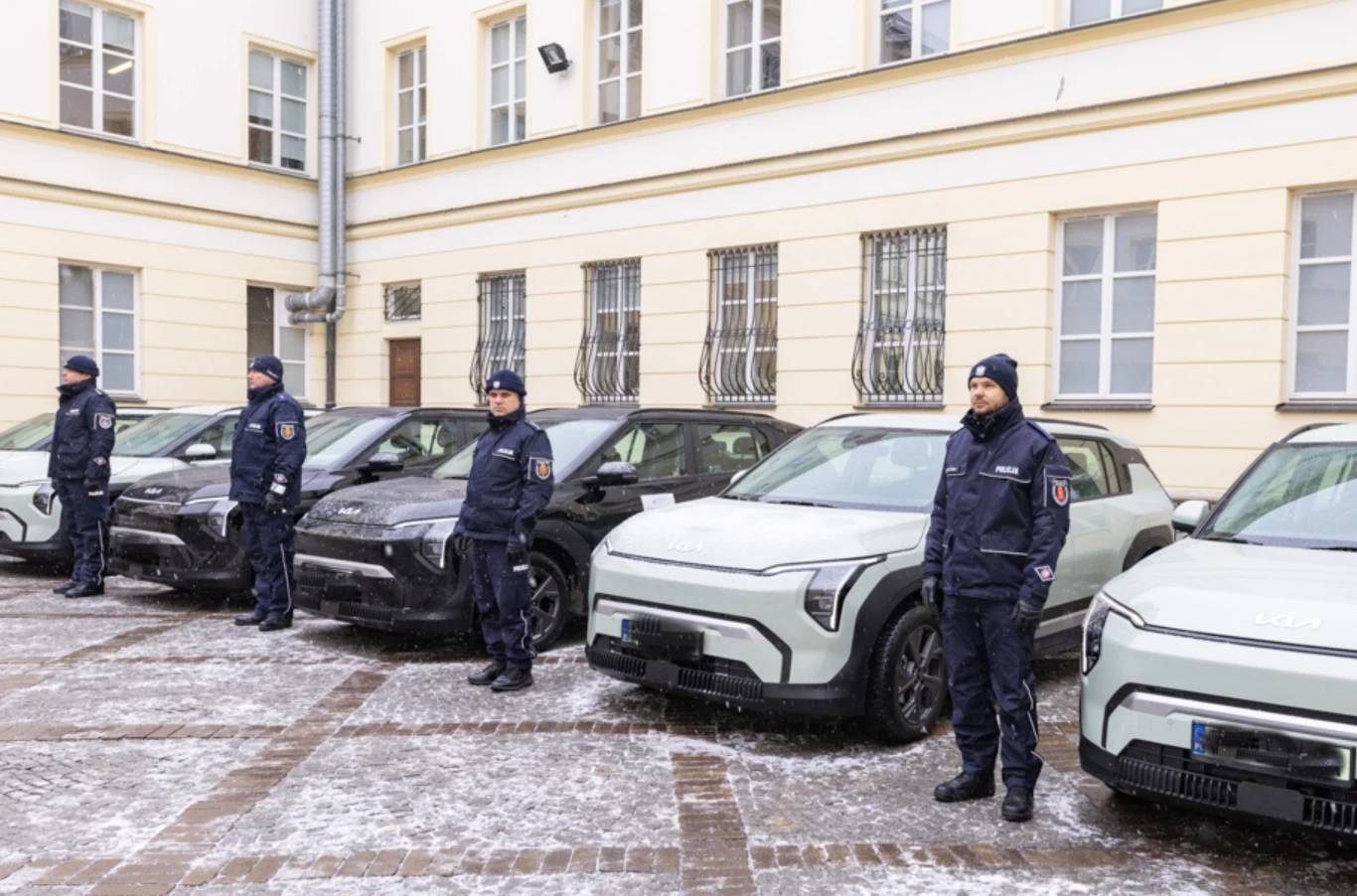 Policjanci w zimowych mundurach stoją w szeregu obok nowoczesnych radiowozów na dziedzińcu przed budynkiem urzędu, na ziemi leży cienka warstwa śniegu.